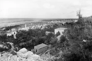 View over the village of Lapithos towards the sea, with a tall tower and dense trees in the valley