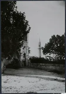 Stone building with an arched gateway and iron gate at Lapithos, a large tree beside it and a slender tower behind