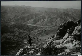 Man standing on a rocky outcrop above a wide valley of bare hills, seen from behind.