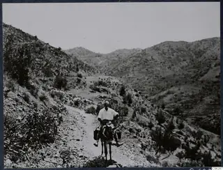 A man rides a donkey down a stony mountain track between dry, scrubby hills.