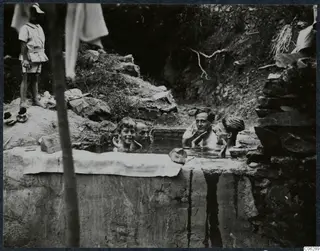 Four people bathing in a stone-lined cistern at Machairas, with a boy standing on the bank.