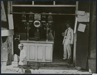 Glass juice dispensers with brass taps on a shop counter, a man in a light suit standing in the doorway