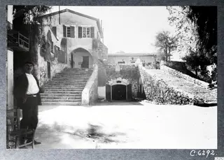 Courtyard with a broad stone staircase, an arched doorway and a man standing at left beside a chair