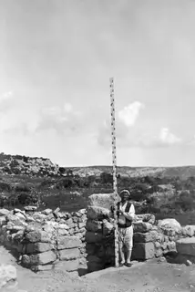 Man holding a tall graduated measuring rod beside a stone wall at Nitovikla