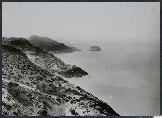 Rocky coastline with scrub-covered slopes falling to the sea, with a small rocky islet offshore.