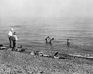 Two men on a pebble beach watch three bathers wading in the sea at Vouni