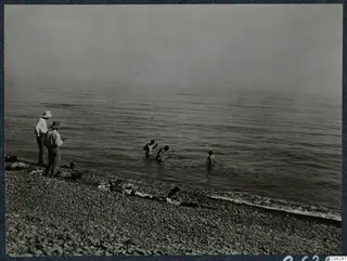 Two men stand on a pebble beach watching three young figures wading in the calm sea