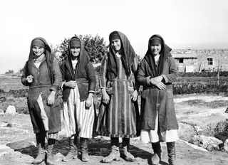 Four young women in headscarves and long coats stand in a row outside a stone building at Vouni.