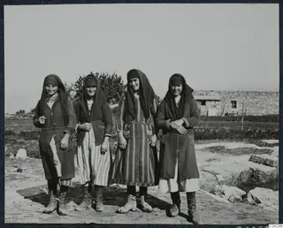 Four young women in headscarves and long coats stand in a row outside, with a stone building behind them, at Vouni.