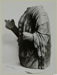 Headless draped figure fragment with one arm raised and the other bent across the body, shown against a pale backdrop