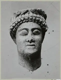 Limestone head of a figure with a banded headdress and wide almond-shaped eyes, photographed against a plain grey background.