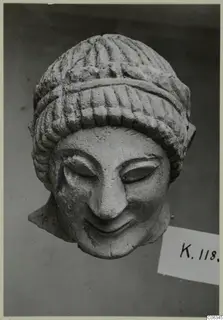 Limestone head of a smiling figure with grooved hair, shown against a dark grey backdrop