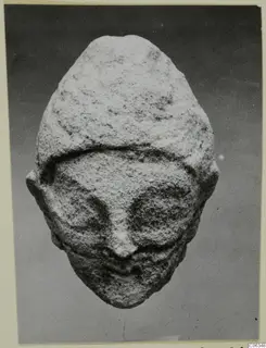 Limestone head from a statuette from Kition, frontal view with prominent nose and deep eye hollows, against a grey backdrop.
