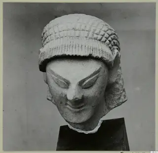 Limestone head statuette from Kition, with a patterned cap and a slight smile, shown on a dark pedestal