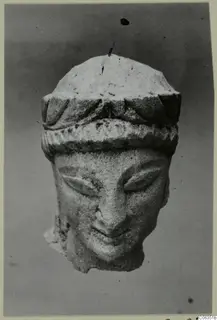 Carved limestone head with a banded headdress and downcast eyes, photographed against a grey background.