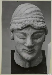 Limestone head of a figure with lowered eyelids and banded hair, set on a dark square base and photographed from the front.