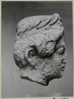 Limestone head fragment with grooved wavy hair and a spiral curl, shown against a plain grey backdrop.