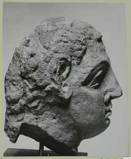 Carved stone head from Mersinaki, shown in profile facing right on a wooden stand.