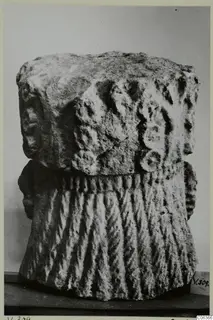 Carved limestone fragment with rounded relief on its upper part and vertical ridges below, shown on a table against a grey backdrop.