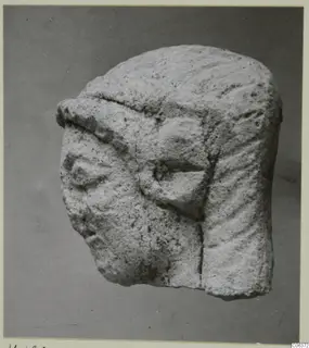 Limestone head fragment from Vouni with carved eyes and grooved hair, shown against a grey backdrop