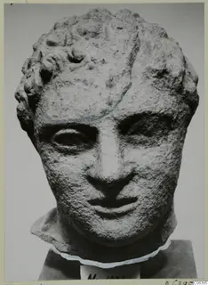 Stone head with curly hair and a crack across the forehead, shown on a dark block base at Mersinaki