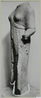 Headless limestone statue of a standing figure in a long garment, photographed against a grey backdrop