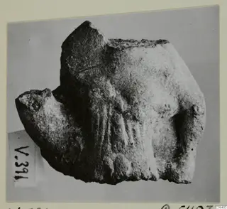 Broken figurine fragment from Vouni with a rough, worn surface, shown on a plain grey backdrop.