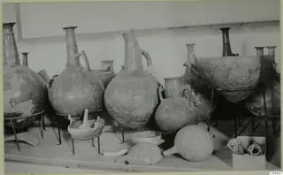 Several large jugs, a bowl and a round flask with patterned surfaces, displayed on metal stands in a museum room.