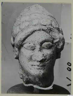 Limestone head from Vouni with a layered headdress, shown front-on on a round wooden stand.