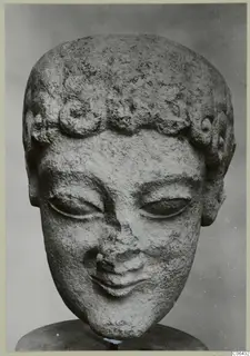 Limestone head of a statue with curled hair and downcast eyes, shown on a dark stand against a grey backdrop.