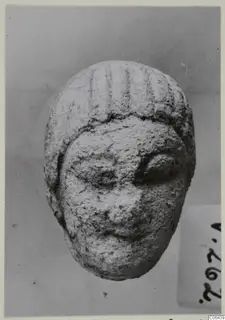 Small limestone head with incised lines on the hair, photographed from the front against a grey backdrop.