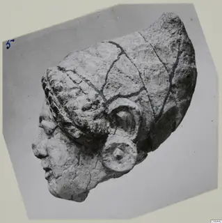 Limestone head fragment in profile facing left, with a ridged headdress and a rounded ear with a disc below it, photographed on grey card.
