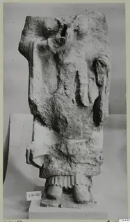 Limestone fragment of a standing figure with the head missing, set on a plinth in front of a grey backdrop