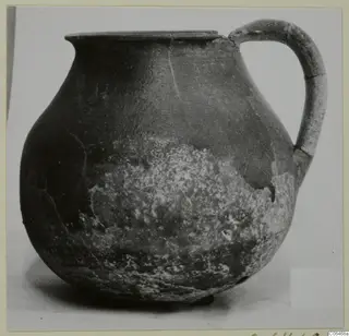 Coarse ware jug with a rounded body, flat rim and strap handle, photographed against a pale background.