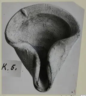 Terracotta lamp with a shallow open bowl and a long spout, photographed from above on a grey mount.