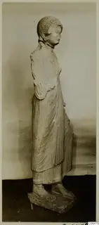 Limestone statue of a standing woman from Vouni, shown in profile in a long pleated garment on a rectangular base.