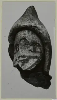 Small terracotta head fragment with a pointed headdress, shown against a plain grey backdrop