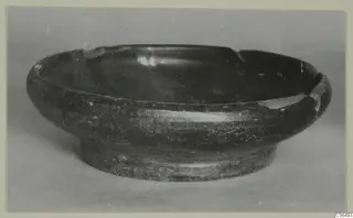 Dark bowl with a ring foot and chipped rim, photographed in side view against a plain grey background