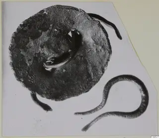 Corroded metal disc with a looped fitting and curved metal fragments beside it
