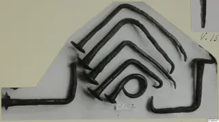 Several dark, corroded metal nails with flat heads and bent shanks, laid out on a grey card with a number label V.103.