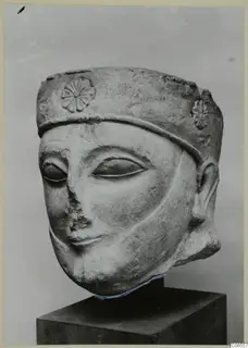 Limestone head sculpture with a tall cylindrical headdress carved with rosettes, shown on a dark pedestal