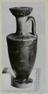 Black glazed ware jug with a flaring rim, a single handle and a ring foot, photographed on a sheet of paper.