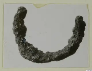 Corroded bronze object curved into an open arc, from Vouni, photographed on a card mount