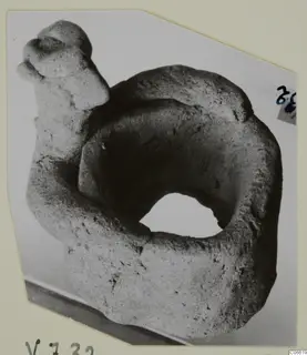 Rough grey clay object with a thick ring-shaped body and a head-like knob on a raised arm, shown on a card mount
