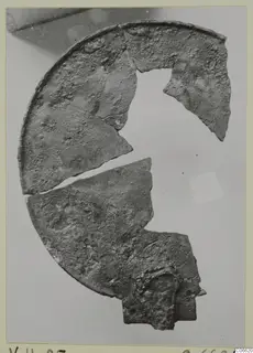 Fragments of a corroded metal disc with a raised rim, from Vouni T. 1-16, shown on a pale backdrop