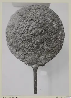Metal mirror from Vouni: a round disc with a long handle, its surface heavily encrusted with corrosion.