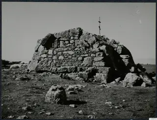 Built tomb of large dressed stone blocks with a pole on top, standing on open, stony ground