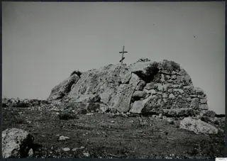 Built tomb with a stone-walled section and a large rock outcrop, topped by a wooden cross on a rocky hillside.