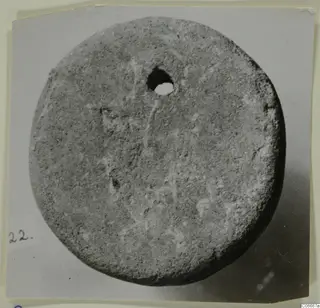 Flat round grey disc with a hole pierced near its upper edge, photographed on a card.