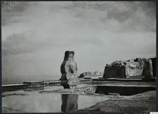 Tall worked stone pillar standing among ruined walls and paved slabs at Vouni palace, with a pale horizon behind.